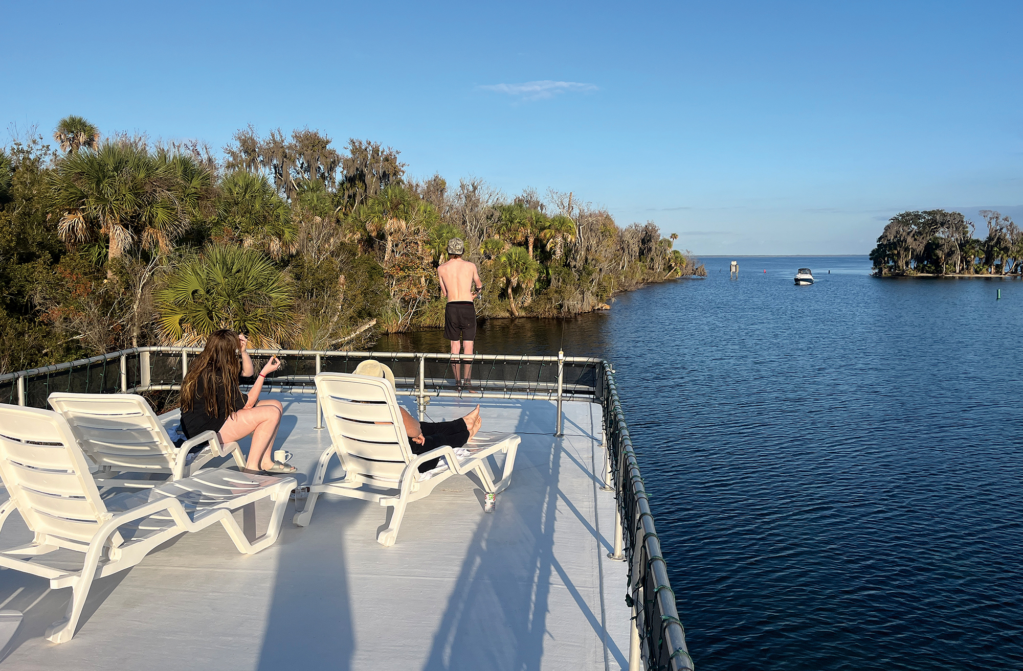 A high-angle shot from the spacious, white upper sun deck of the houseboat River Time as it navigates a scenic waterway lined with lush palmetto and cypress trees. In the foreground, white plastic lounge chairs face the water, where family members are relaxing and looking out at the scenery. A shirtless man stands near the safety netting at the edge, gazing at the calm, dark blue river. In the distance, a small boat cruises near a tree-covered island under a clear blue sky.