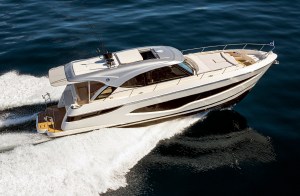 An elevated, high-angle aerial shot of a sleek white Riviera luxury sports express motor yacht cruising rapidly through deep blue open water. The modern vessel features sweeping dark-tinted hull windows, a large sunpad on the bow deck, and a hardtop roof with an open sunroof. The boat carves smoothly through the sea, casting a crisp reflection on the water's surface to starboard while leaving a powerful, foaming white wake trailing behind its teak-planked swim platform.