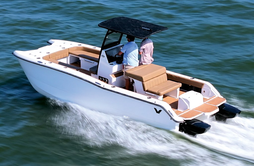 An aerial, high-angle three-quarter shot of a white center-console power catamaran cruising through green water, leaving a foaming white wake behind its twin hulls. Two men are on board, with the driver at the helm station beneath a black T-top canopy. The boat features tan upholstered seating, including an open bow lounge area and a rear-facing cockpit bench. The aft deck is finished with a light brown faux-teak swim platform extension flanking twin black outboard engines.