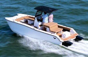 An aerial, high-angle three-quarter shot of a white center-console power catamaran cruising through green water, leaving a foaming white wake behind its twin hulls. Two men are on board, with the driver at the helm station beneath a black T-top canopy. The boat features tan upholstered seating, including an open bow lounge area and a rear-facing cockpit bench. The aft deck is finished with a light brown faux-teak swim platform extension flanking twin black outboard engines.