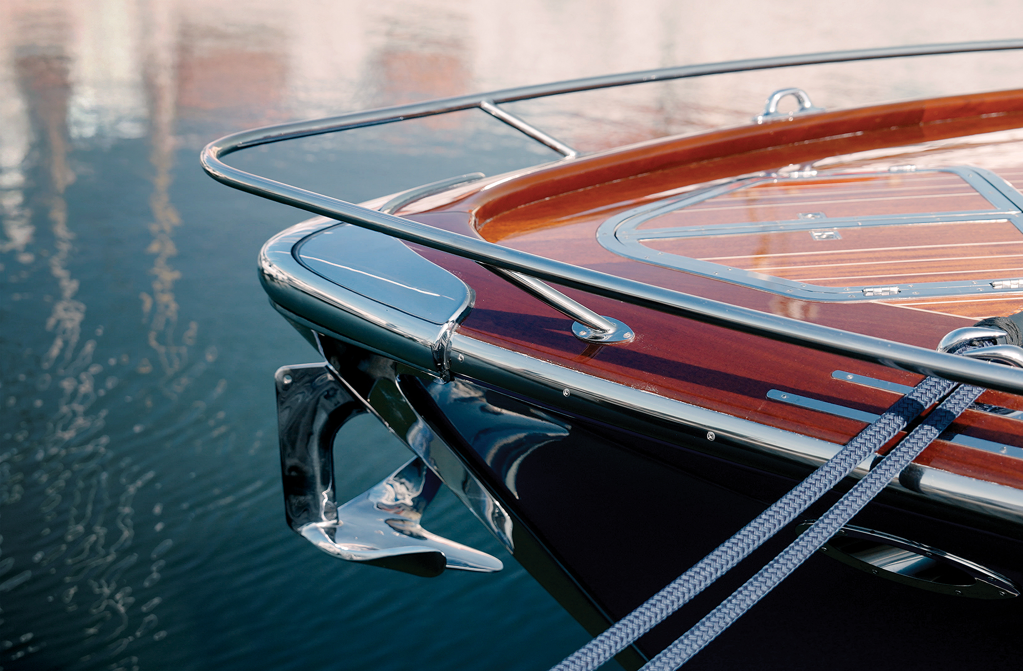 A close-up, high-angle view of the bow of a luxury motor yacht moored on calm water. The bow features highly polished, varnished mahogany wood decking with a stainless steel bow rail, a prominent chrome anchor mounted at the stem, and two braided blue dock lines stretching diagonally across the frame.