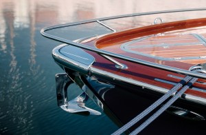A close-up, high-angle view of the bow of a luxury motor yacht moored on calm water. The bow features highly polished, varnished mahogany wood decking with a stainless steel bow rail, a prominent chrome anchor mounted at the stem, and two braided blue dock lines stretching diagonally across the frame.