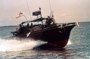 A vintage, low-angle historical photograph of a military Patrol Boat River (PBR) cruising through choppy river water. The dark, camouflage-painted combat vessel features a rounded black hull with a red bottom, kicking up a white water spray from its hull. Soldiers wearing military helmets and gear are visible on board, with a crewman stationed at the helm beneath a black canvas canopy structure equipped with antennas and a radar unit. An American flag flies prominently from the stern, and a machine gun mount is positioned at the back of the vessel under an overcast sky.