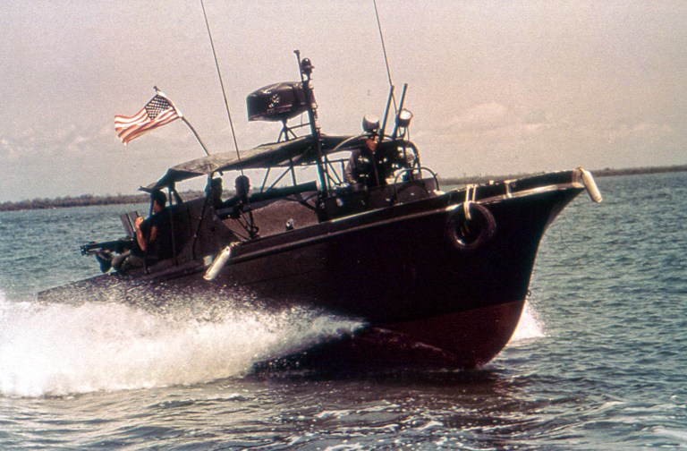 Blank old photo isolated on white A vintage, low-angle historical photograph of a military Patrol Boat River (PBR) cruising through choppy river water. The dark, camouflage-painted combat vessel features a rounded black hull with a red bottom, kicking up a white water spray from its hull. Soldiers wearing military helmets and gear are visible on board, with a crewman stationed at the helm beneath a black canvas canopy structure equipped with antennas and a radar unit. An American flag flies prominently from the stern, and a machine gun mount is positioned at the back of the vessel under an overcast sky.