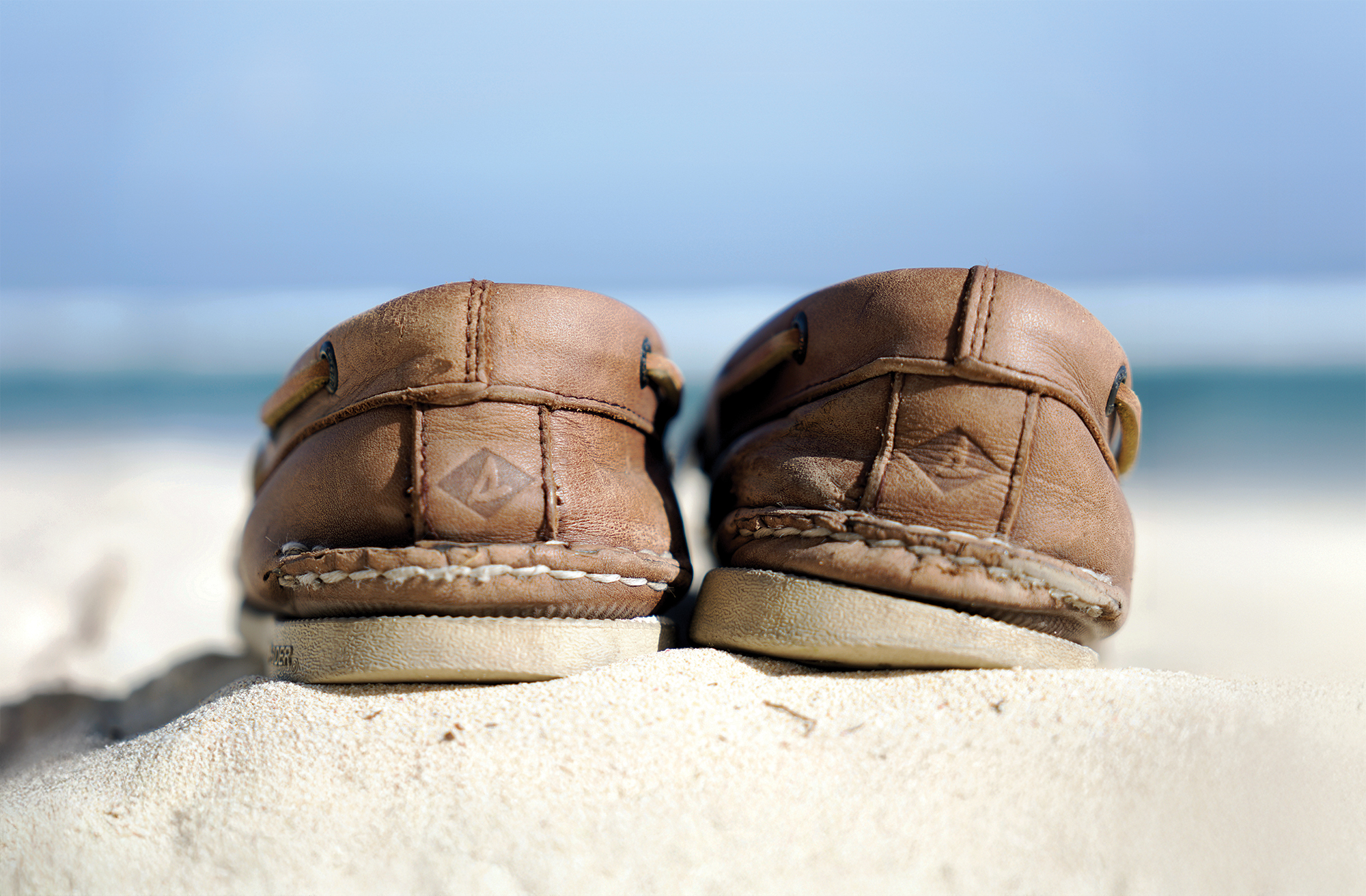 A close-up, low-angle shot from behind a pair of well-worn, brown leather Sperry Top-Sider boat shoes sitting on a white sandy beach. The focus centers on the stitched heels, each featuring the embossed diamond Sperry logo on the leather. The shoes rest on a small sand dune, with a softly blurred background showing turquoise ocean water under a pale blue sky.