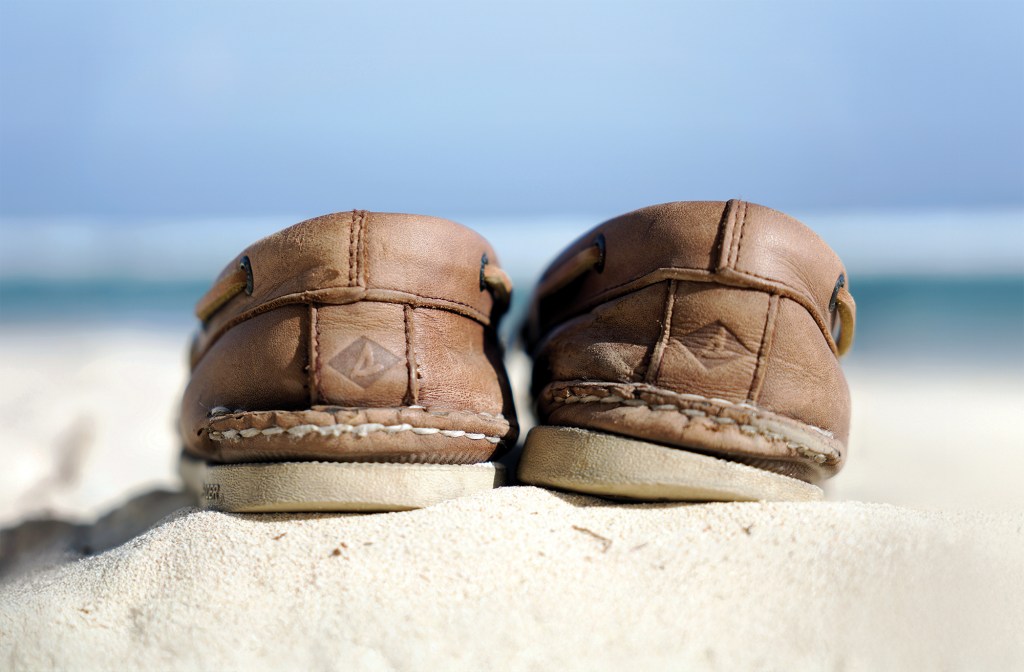 A close-up, low-angle shot from behind a pair of well-worn, brown leather Sperry Top-Sider boat shoes sitting on a white sandy beach. The focus centers on the stitched heels, each featuring the embossed diamond Sperry logo on the leather. The shoes rest on a small sand dune, with a softly blurred background showing turquoise ocean water under a pale blue sky.