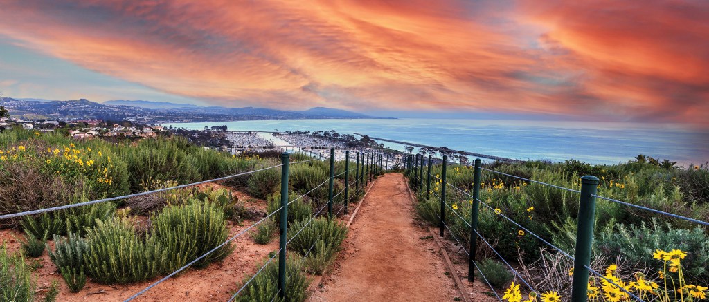 A panoramic, high-angle view looking down a fenced dirt hiking trail on the Dana Point Headlands, leading toward the harbor and open ocean under a dramatic orange sunset sky.