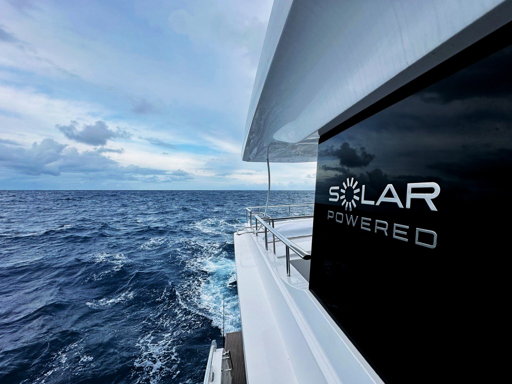 A view looking out at the open blue ocean from the deck of a yacht, featuring a glossy black panel on the side of the superstructure with a silver "Solar Powered" emblem.