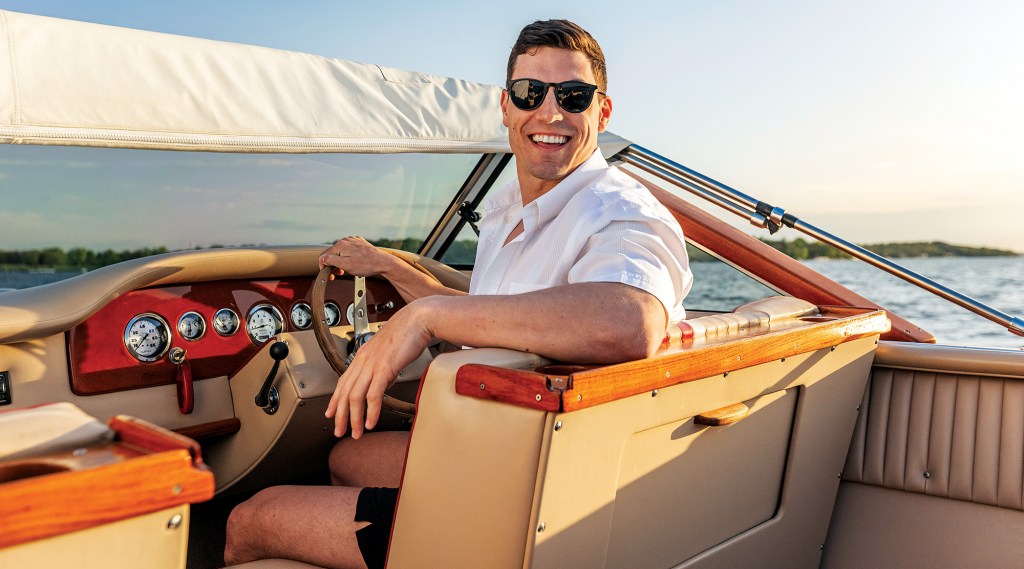 A smiling man wearing sunglasses and a white shirt sitting at the helm of a classic wooden boat on a sunny lake.
