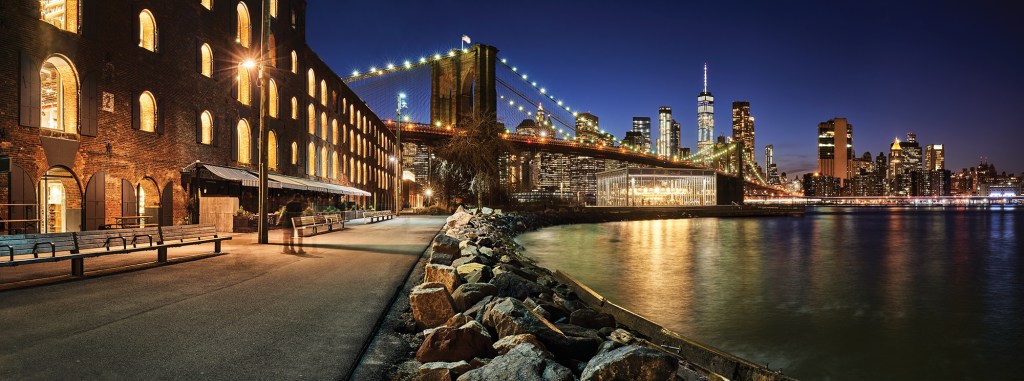 A panoramic night view from a Brooklyn waterfront walkway, featuring an illuminated historic brick building, the lit spans of the Brooklyn Bridge, and the glowing Manhattan skyline reflecting on the East River.