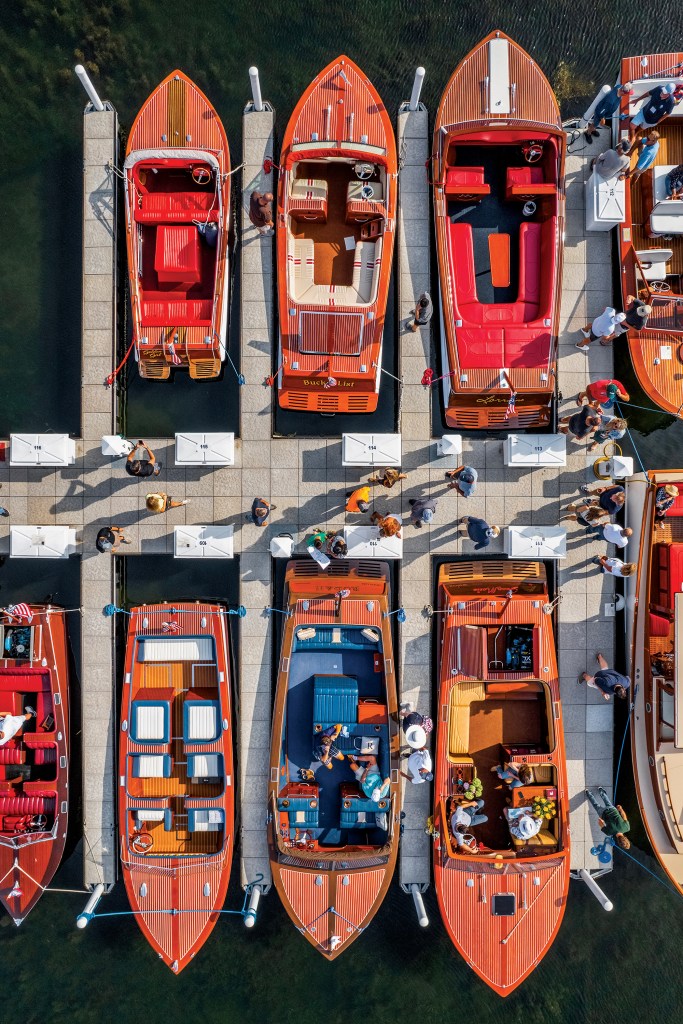 An overhead aerial view of several classic mahogany wooden boats docked along floating piers at a marina with people walking nearby.