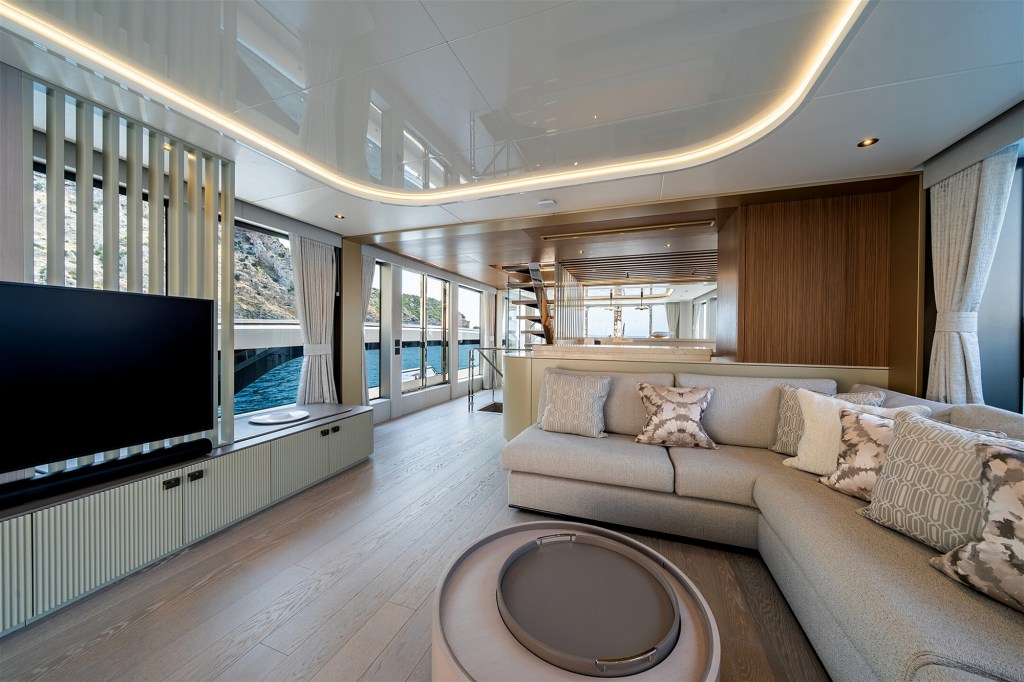 An interior view of the main deck salon inside the Sunseeker 82 Ocean yacht, showcasing a luxurious and modern open-concept design. On the right, a large, plush light grey L-shaped sectional sofa with patterned accent pillows faces a round coffee table with a grey tray insert resting on light wood flooring. To the left, a large flat-screen television is positioned over a fluted media console cabinet next to floor-to-ceiling windows that reveal water and a rocky cliffside outside. The space is illuminated by a glossy white ceiling accented with a curved, recessed track of warm LED strip lighting.