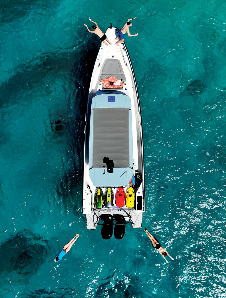 An aerial overhead view of an Axopar 37 Sun Top boat anchored in vibrant turquoise water, with passengers diving into the ocean from the bow while others swim nearby in the crystal-clear sea.