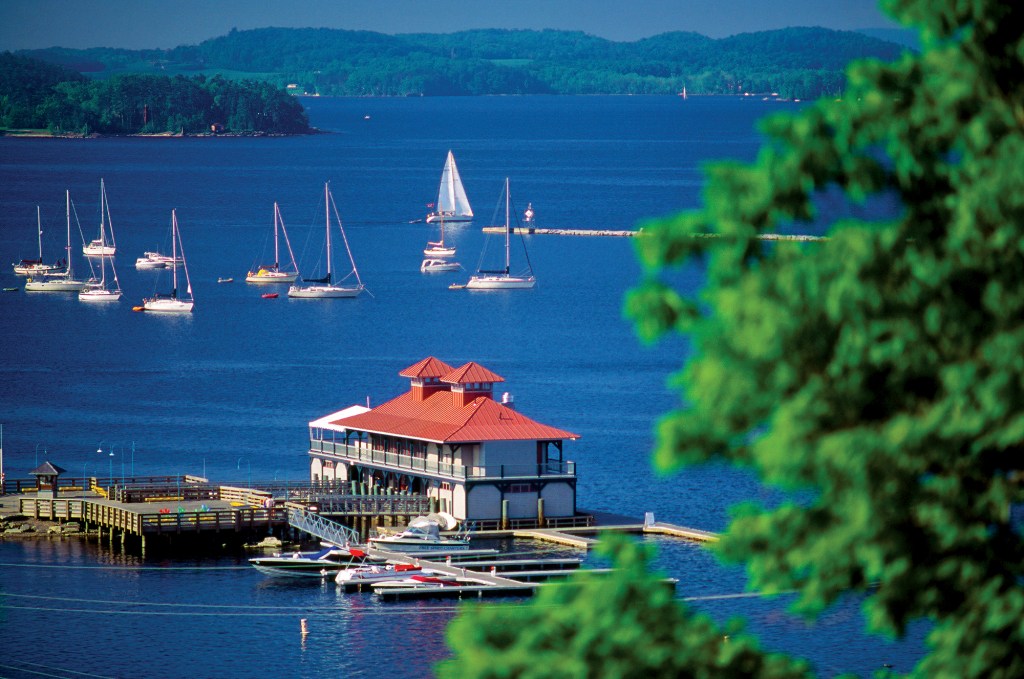 A high-angle view of a red-roofed boathouse and pier on Lake Champlain, with numerous sailboats anchored on the blue water and forested hills rising in the distance.