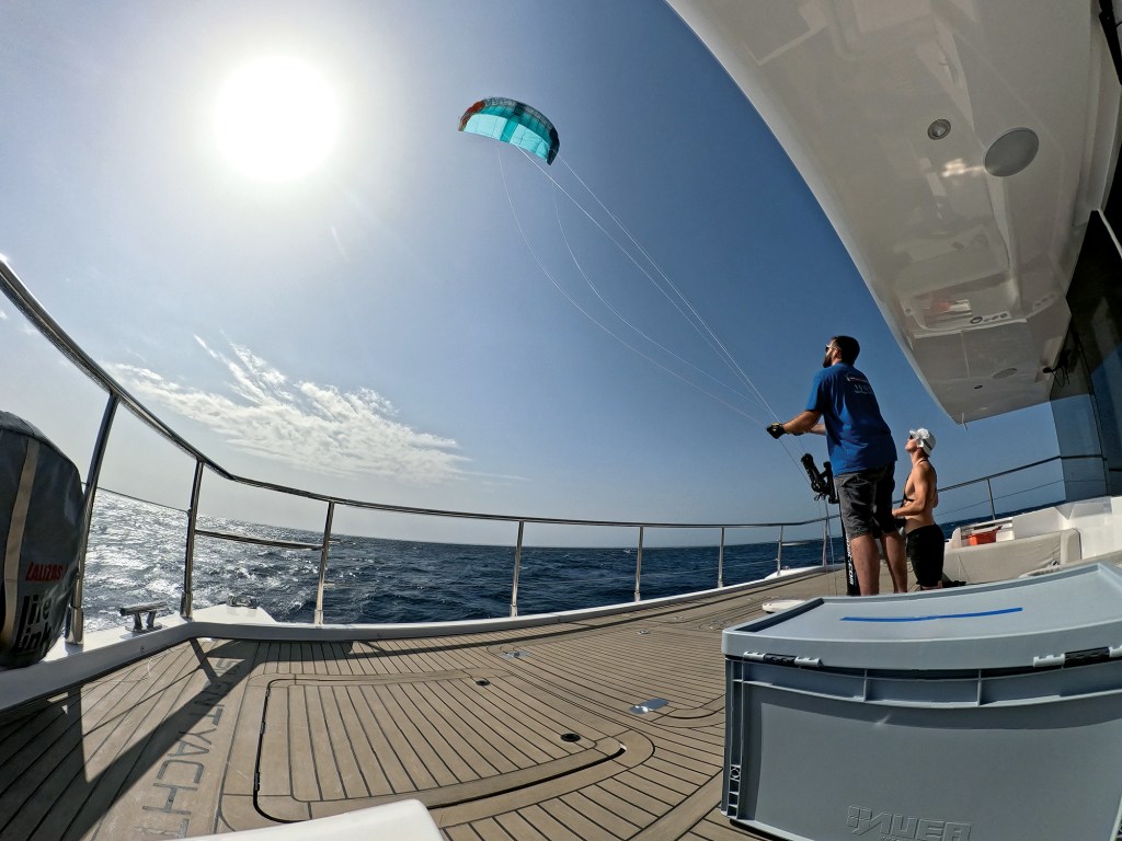 A low-angle, wide shot from the deck of a yacht showing crew members launching an automated green-and-blue propulsion kite into the sky on a bright, sunny day at sea.