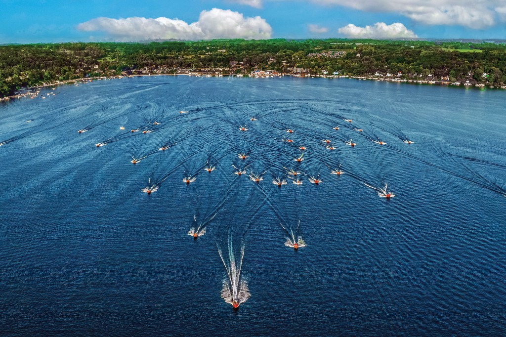 An aerial view of dozens of wooden motorboats cruising in formation across a large blue lake toward a lush green shoreline.
