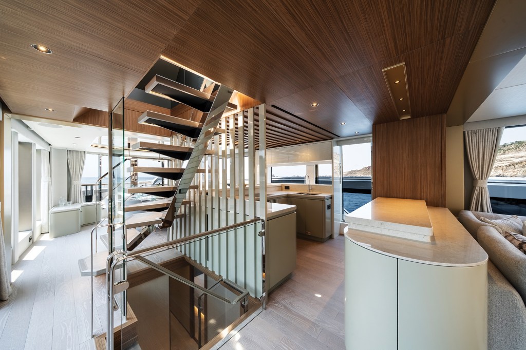 An interior view of the main deck on the Sunseeker 82 Ocean yacht, highlighting a central open-riser architectural staircase with glass railings and wood treads leading to the upper deck. To the right of the staircase, a modern galley kitchen features light beige cabinetry, solid surface countertops, and a sink beneath large windows overlooking the sea. The space is anchored by warm wood-veneer ceiling panels, light oak flooring, and a curved service counter bar in the foreground.