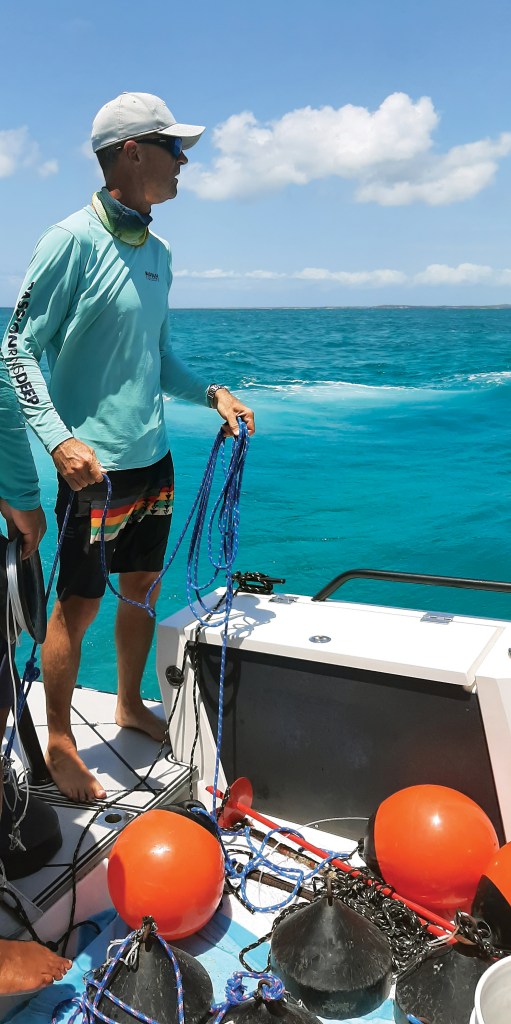 A man standing on the stern deck of a boat in turquoise water, holding blue coiled rope near orange buoys and fishing equipment while looking out across the sea.