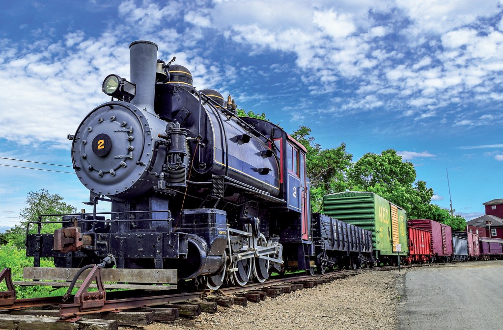 A low-angle shot of a vintage black steam locomotive, marked with the number 2, pulling a line of colorful freight cars on a railroad track under a bright blue sky with white clouds.