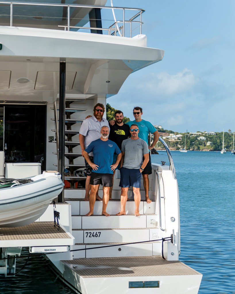 Five crew members posing together on the wide stern steps of a large white catamaran yacht docked in calm coastal waters, with a lush tropical coastline in the background.