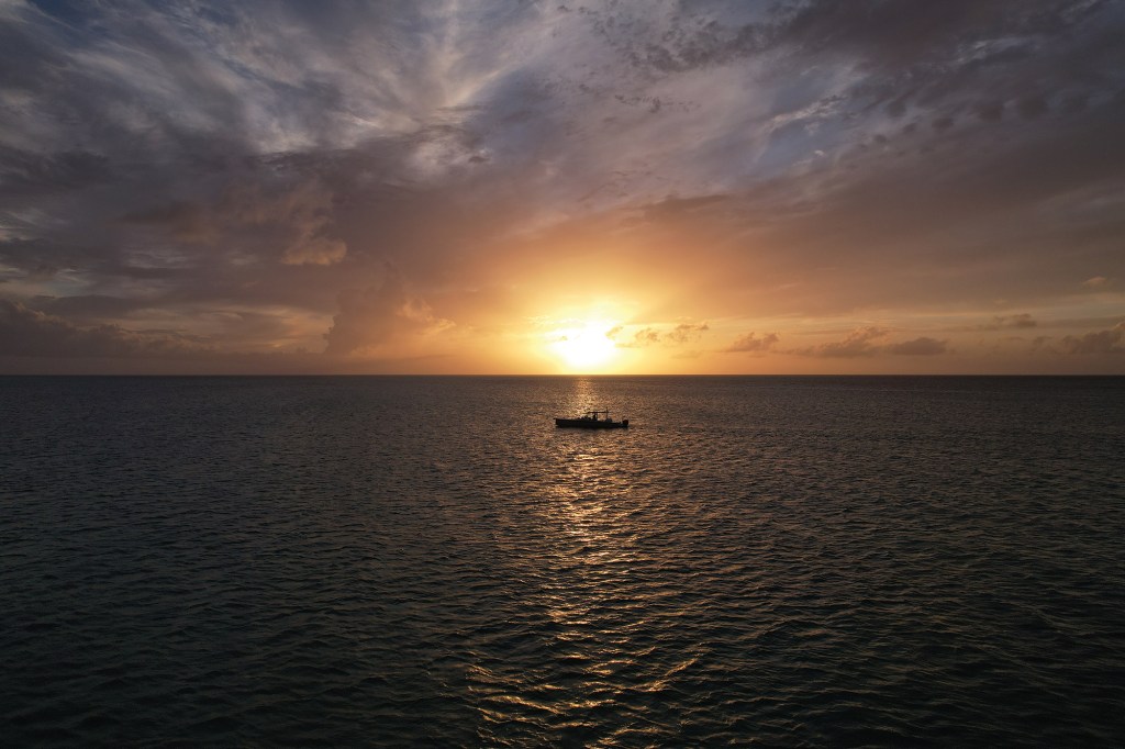 A wide aerial view of a single boat silhouetted against a golden sunset over the calm open ocean.