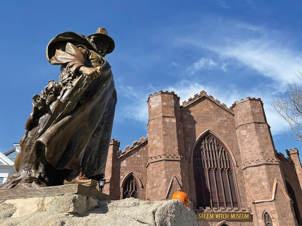 A low-angle shot of the bronze Roger Conant statue standing prominently in the foreground, with the stone facade and Gothic-style towers of the Salem Witch Museum visible behind it under a blue sky.