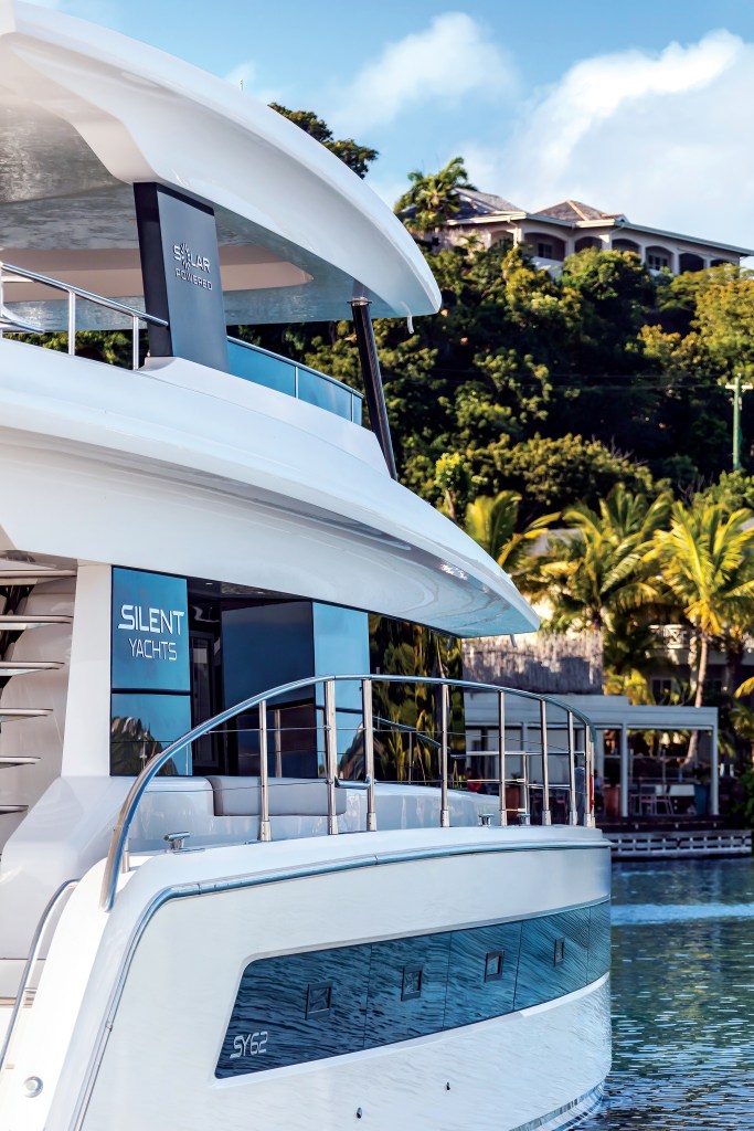 A close-up profile view of the multi-level white superstructure of a Silent Yachts SY62 catamaran, featuring "Silent Yachts" and "Solar Powered" branding, with a lush tropical hillside in the background.