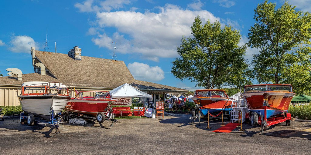 Several classic wooden boats displayed on trailers outdoors in front of a Streblow Custom Boats event setup.