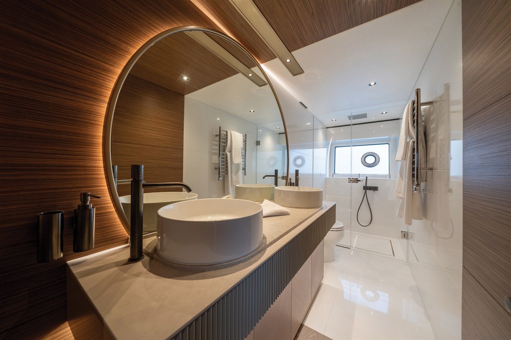 A contemporary en suite bathroom inside the master stateroom of the Sunseeker 82 Ocean yacht, designed with high-end, spa-like finishes. In the foreground, a modern vanity with a beige solid-surface countertop holds two white vessel sinks paired with dark bronze faucets beneath a large, circular backlit mirror. The left wall is finished in horizontal wood grain paneling, contrasting with a polished white marble floor that flows back toward a glass-enclosed walk-in shower featuring a porthole window.