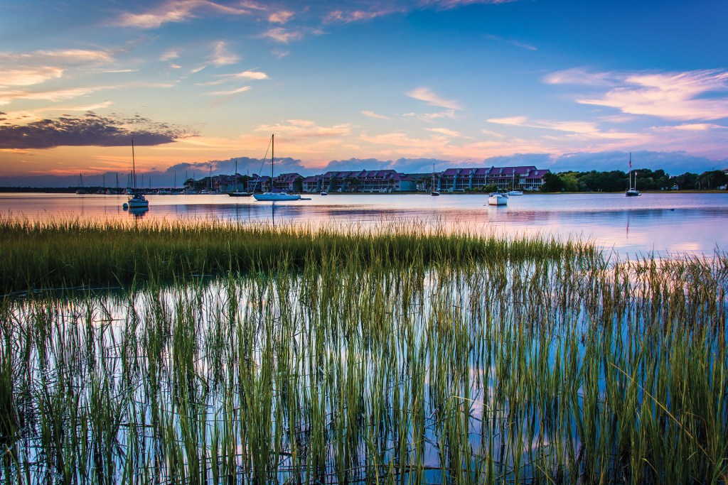 A scenic view looking across a salt marsh with tall green cordgrass in the foreground, out toward a calm harbor where sailboats are anchored in front of a row of coastal condominiums during a pink and blue sunset.
