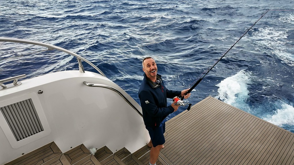 A high-angle view of a smiling man holding a fishing rod while standing on the wide teak stern platform of a catamaran yacht cruising through deep blue open ocean water.