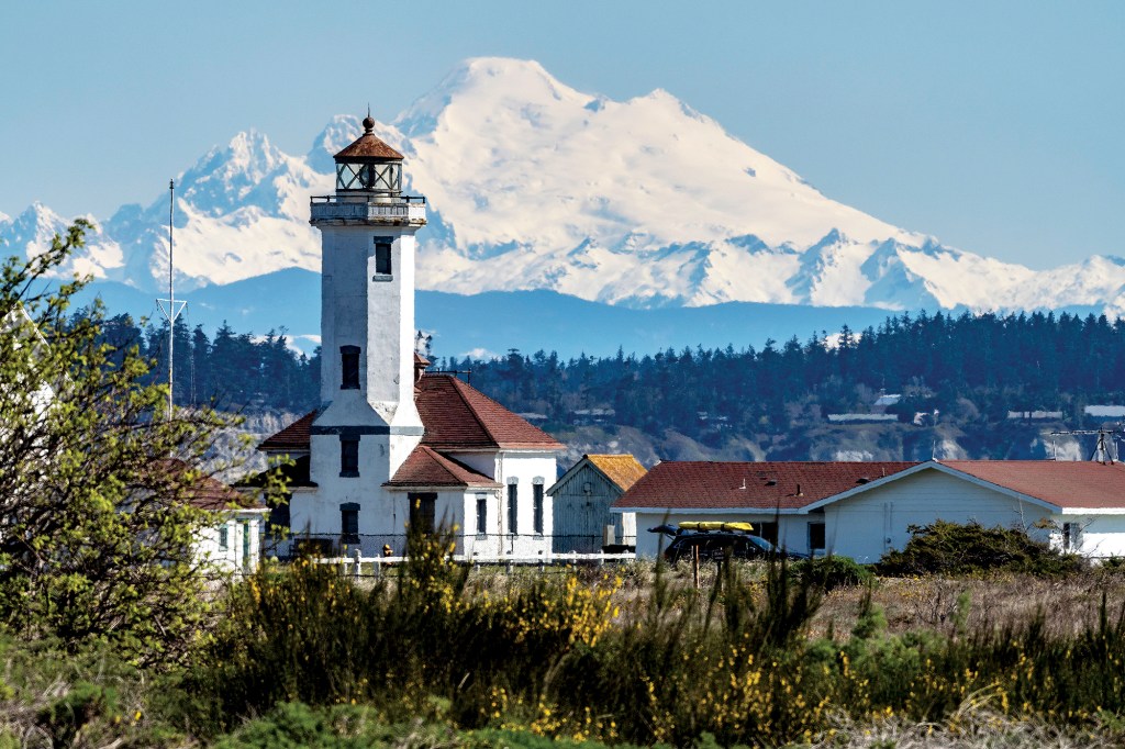 A distance shot of the historic Point Wilson Lighthouse at Fort Worden State Park in Port Townsend, Washington, showing its white octagonal tower and red-roofed buildings against the backdrop of the massive, snow-covered peak of Mount Baker.