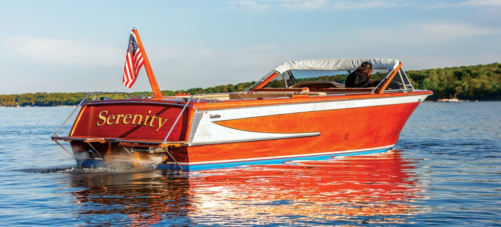 A classic mahogany wooden boat named Serenity navigating calm lake water with an American flag flying from the stern.