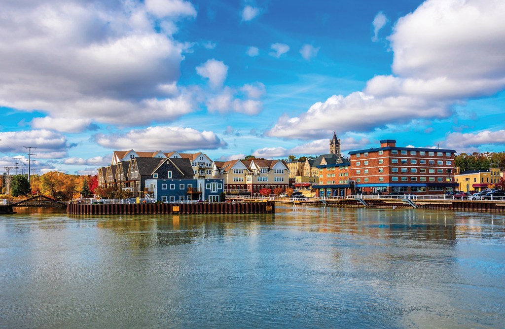 A wide, scenic view looking across the calm waters of the harbor toward the waterfront buildings and historic downtown architecture of Port Washington, Wisconsin, under a blue sky filled with puffy white clouds.
