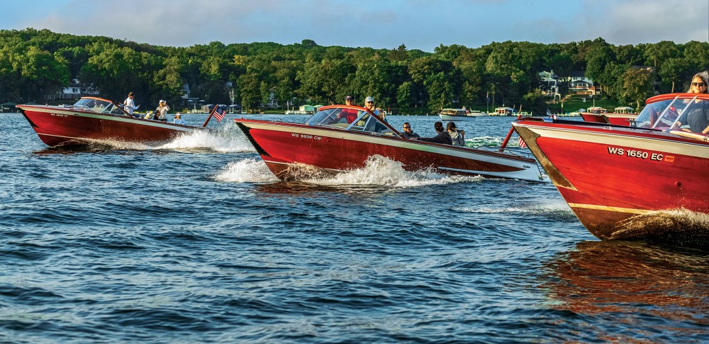 Three wooden runabout boats cruising in formation across a lake near a tree-lined shore.