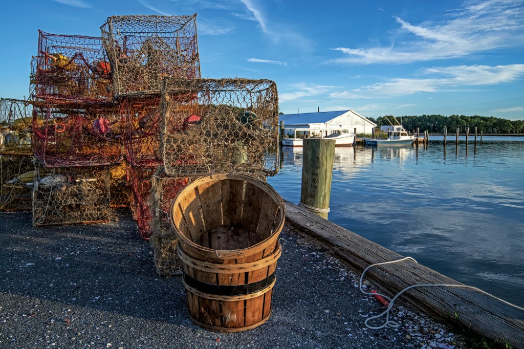 A low-angle shot of a rustic wooden bushel basket and a stack of weathered crab traps on a waterfront pier in St. Michaels, Maryland, with fishing boats and a dock house across the calm water.