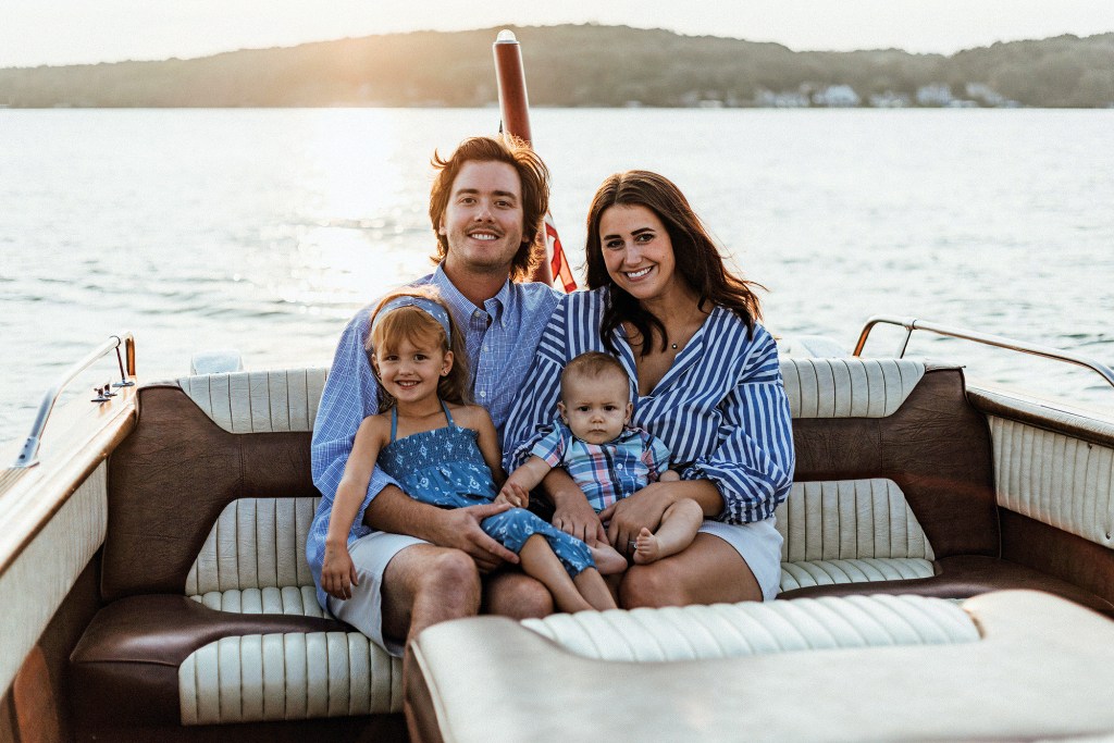 A family of four sitting together on the plush rear bench seat of a wooden boat on a lake at sunset.