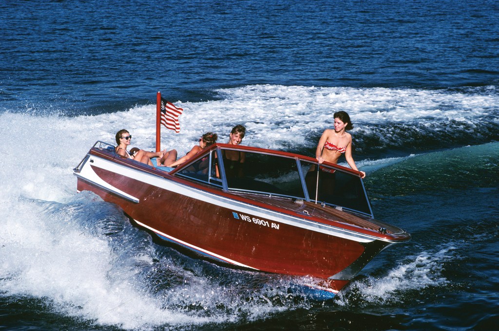 Four women riding in a classic wooden runabout boat speeding across choppy blue water and kicking up white bow spray.