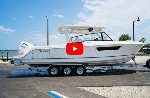 feat-Edgewater 310SC A profile view of a white EdgeWater 310CX powerboat equipped with twin Yamaha outboard engines, resting on a triple-axle Float-On boat trailer on a paved lot, featuring a red video play button overlaid in the center.