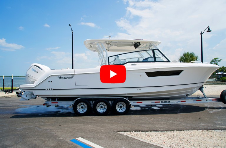 A profile view of a white EdgeWater 310CX powerboat equipped with twin Yamaha outboard engines, resting on a triple-axle Float-On boat trailer on a paved lot, featuring a red video play button overlaid in the center.