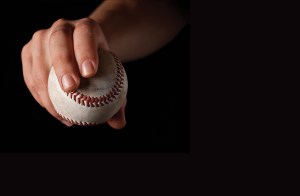 A close-up of a person's hand gripping a scuffed baseball with red stitching against a solid black background.