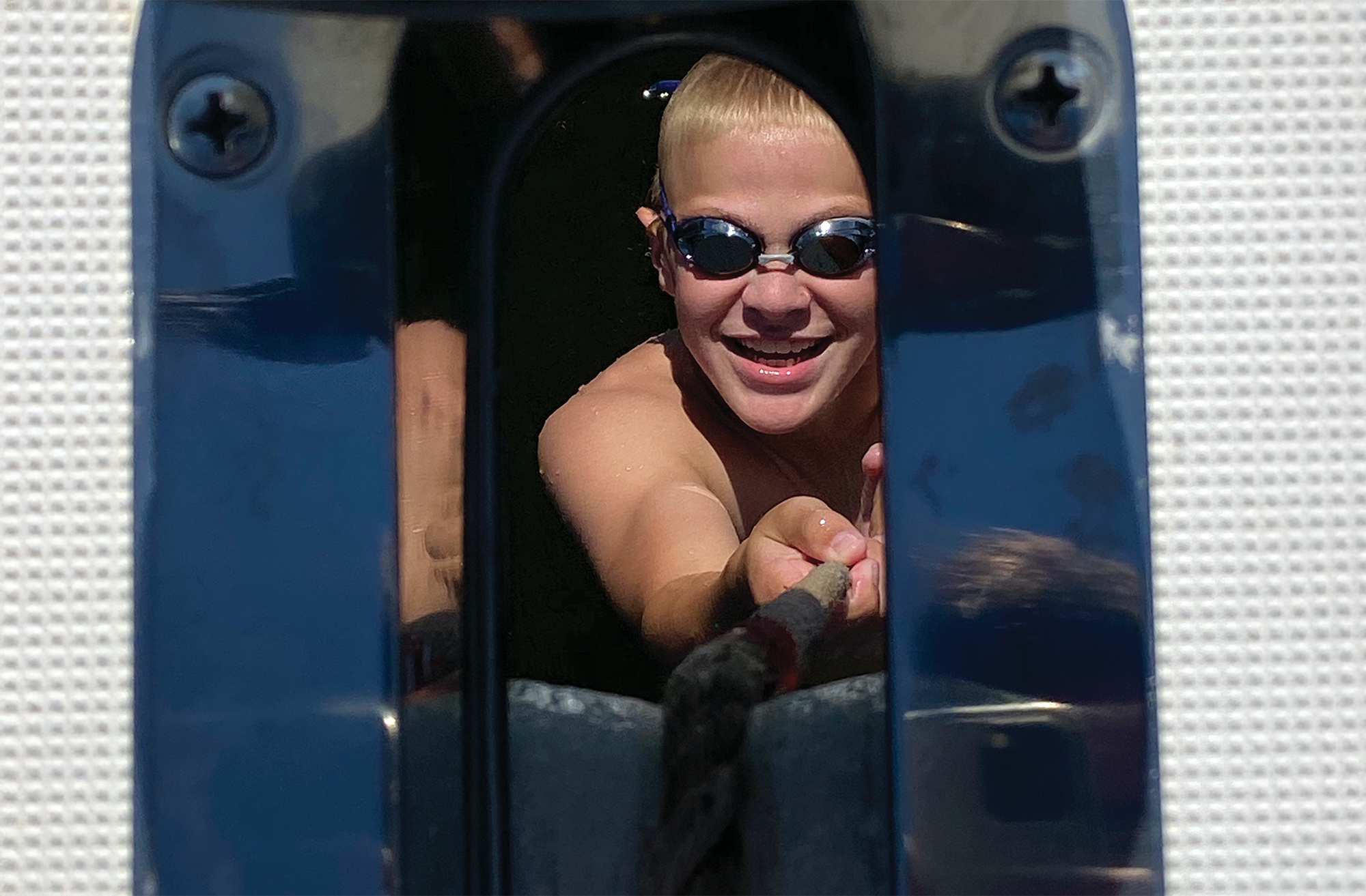 A young boy wearing swim goggles, framed through a boat's bow hawsehole, smiling brightly as he pulls on an anchor rode while swimming in the water.
