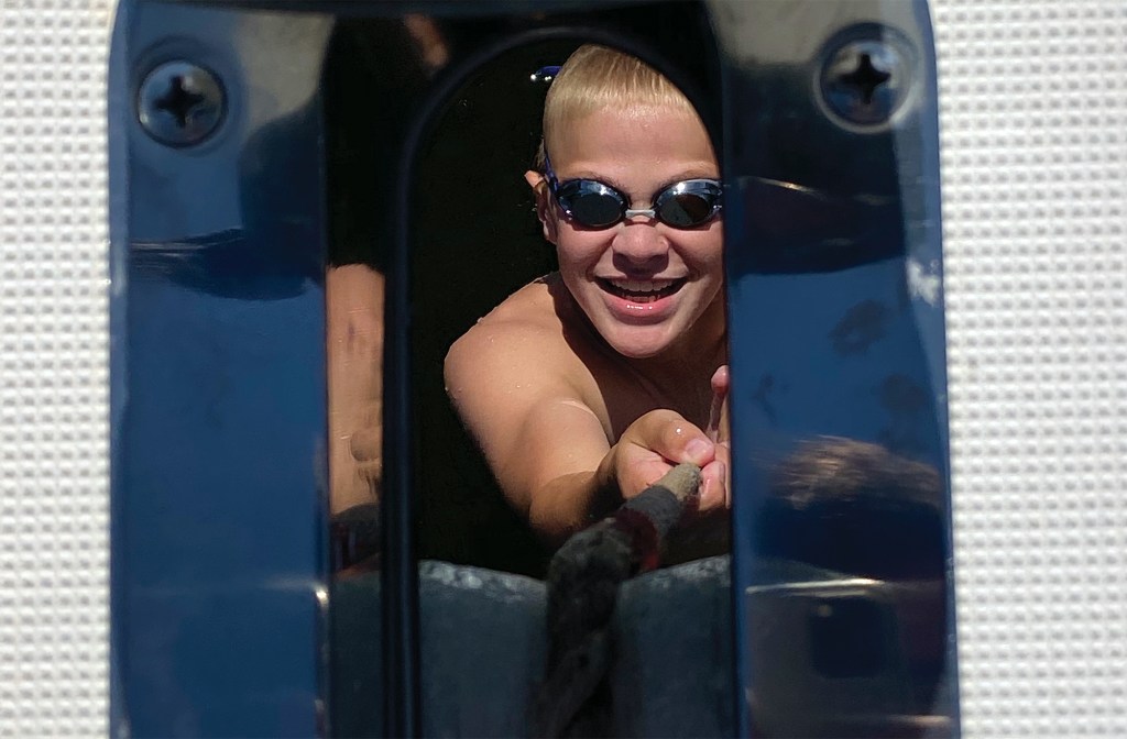 A young boy wearing swim goggles, framed through a boat's bow hawsehole, smiling brightly as he pulls on an anchor rode while swimming in the water.