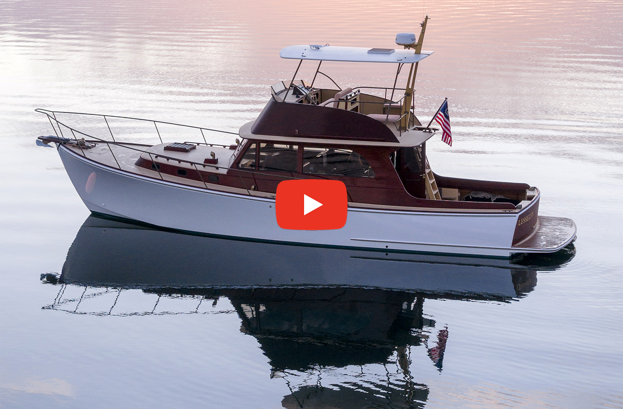 A side-profile view of a classic white Hood 42 LM motor yacht with rich wood accents resting on calm, mirror-like water during sunset, with a red video play button overlaid in the center.