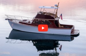 Hood 42 LM powerboat, Lesselyn. A side-profile view of a classic white Hood 42 LM motor yacht with rich wood accents resting on calm, mirror-like water during sunset, with a red video play button overlaid in the center.