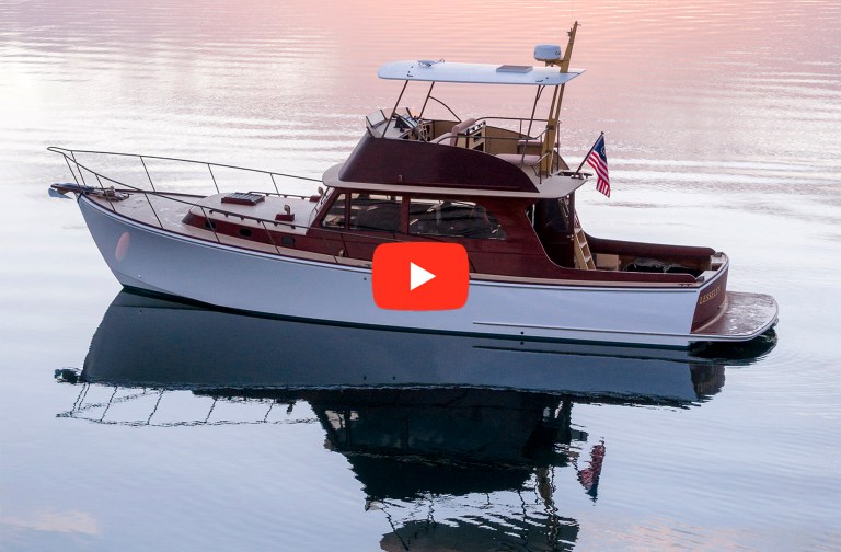 A side-profile view of a classic white Hood 42 LM motor yacht with rich wood accents resting on calm, mirror-like water during sunset, with a red video play button overlaid in the center.