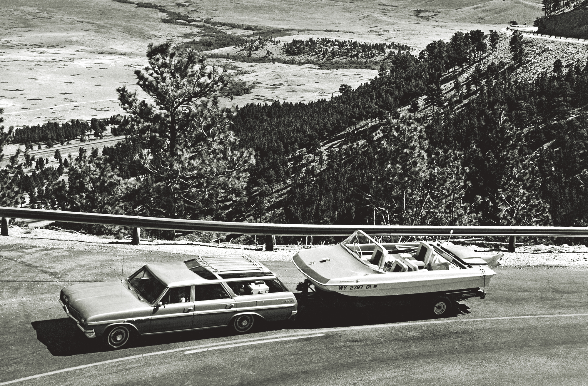 A black and white, high-angle shot of a classic 1960s Buick Sportwagon towing a vintage Evinrude Sweet-16 tri-hull motorboat on a trailer along a scenic mountain highway overlook.