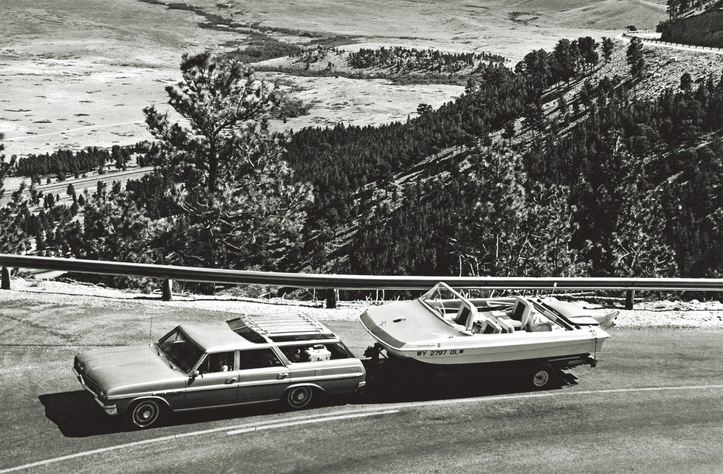 A black and white, high-angle shot of a classic 1960s Buick Sportwagon towing a vintage Evinrude Sweet-16 tri-hull motorboat on a trailer along a scenic mountain highway overlook.