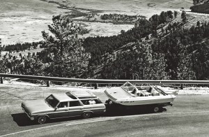A black and white, high-angle shot of a classic 1960s Buick Sportwagon towing a vintage Evinrude Sweet-16 tri-hull motorboat on a trailer along a scenic mountain highway overlook.