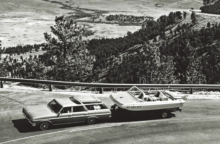 Blank old photo isolated on white A black and white, high-angle shot of a classic 1960s Buick Sportwagon towing a vintage Evinrude Sweet-16 tri-hull motorboat on a trailer along a scenic mountain highway overlook.